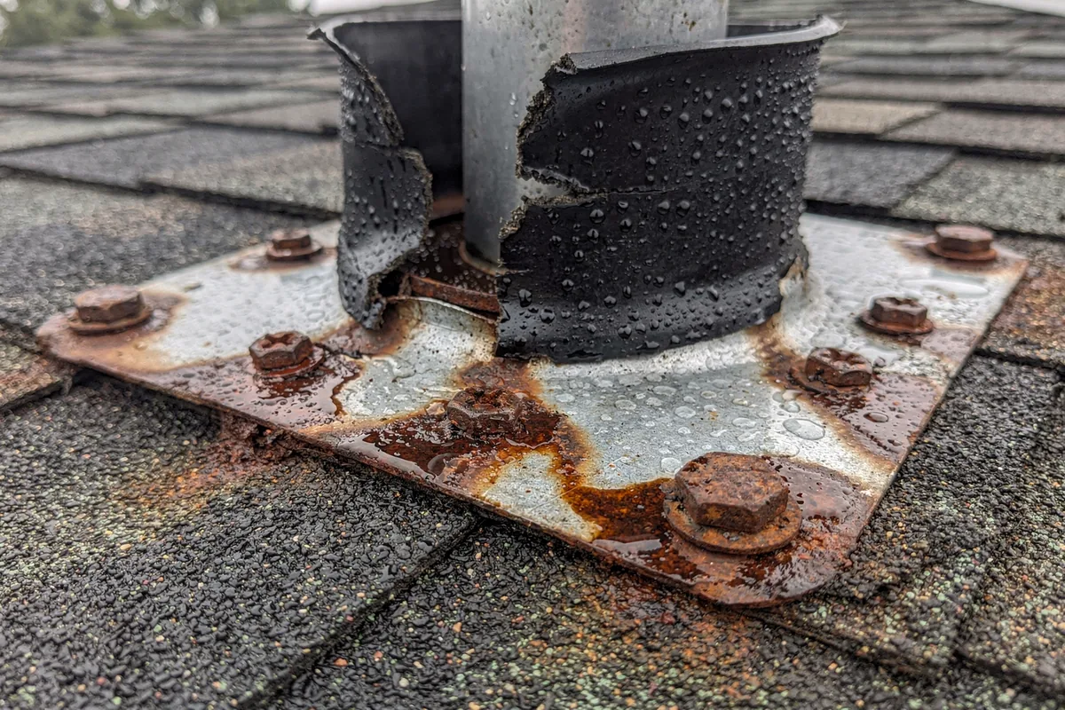 A plumbing vent boot on wet shingles with its collar split open and rust bleeding from the fasteners below it.